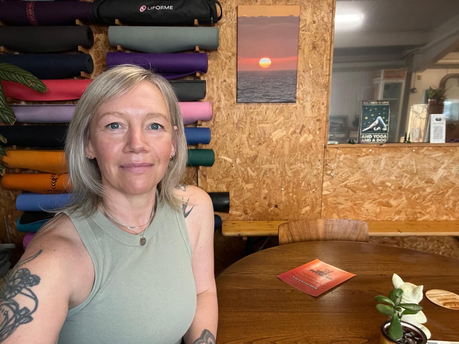 Helen at the table in the studio, with a wall of colourful yoga mats behind her